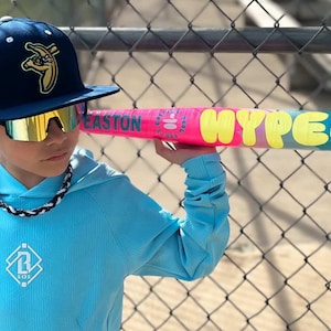 May include: A person wearing a navy blue baseball cap with a banana logo and mirrored sunglasses holds a pink and blue baseball bat. The bat has the words "HYPE FIRE" in yellow. The person is wearing a light blue hoodie.