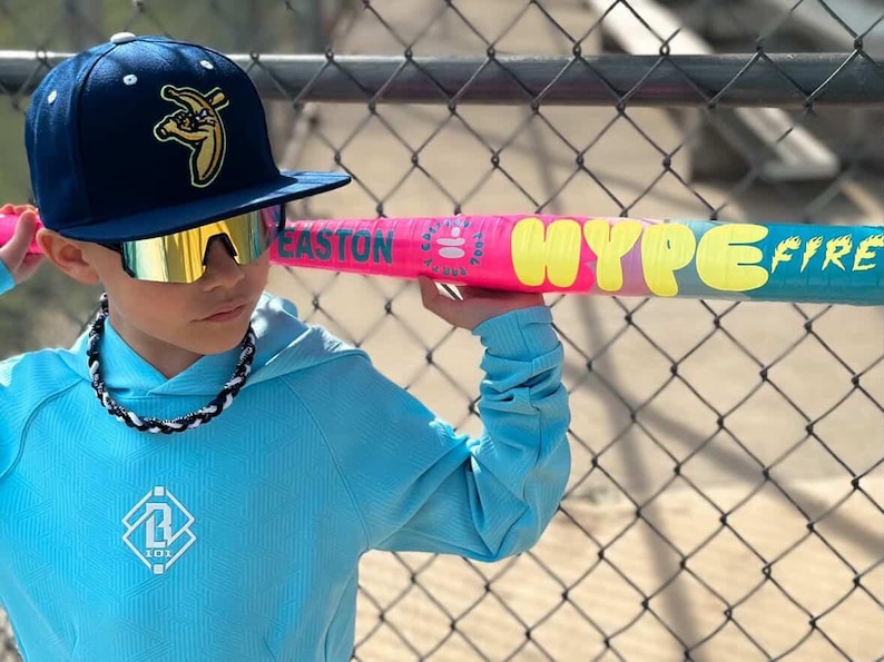May include: A person wearing a navy blue baseball cap with a banana logo and mirrored sunglasses holds a pink and blue baseball bat. The bat has the words "HYPE FIRE" in yellow. The person is wearing a light blue hoodie.