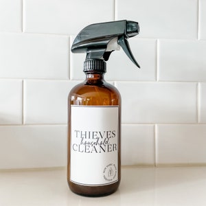 Może przedstawiać: Brązowa szklana butelka z rozpylaczem z czarnym spustem. Butelka ma białą etykietę z napisem "THIEVES household CLEANER" i mniejszą okrągłą etykietę z napisem "NON TOXIC PLANT BASED CLEANING".