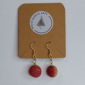 May include: A pair of gold dangle earrings with red and brown clay donut-shaped charms. The earrings are on a brown card with a white circle sticker that says "MALIA GARCIA ARTS" with a fern design.