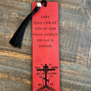 May include: A red leather bookmark with black tassel. The bookmark features the text "LORD JESUS CHRIST SON OF GOD HAVE MERCY ON ME A SINNER" and a cross symbol with Cyrillic text. A white cross is at the top.