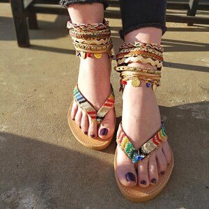 May include: A pair of brown leather sandals with colourful woven straps and gold accents. The sandals are adorned with multiple gold anklets with charms, including evil eye beads.
