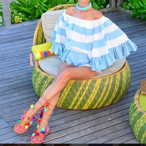 May include: A woman wearing a blue and white striped off-the-shoulder top and colourful pom-pom sandals sits on a woven chair. The chair is made of woven bamboo and has a green and yellow pattern.