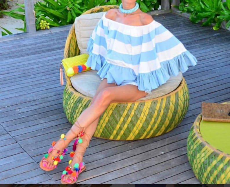 May include: A woman wearing a blue and white striped off-the-shoulder top and colourful pom-pom sandals sits on a woven chair. The chair is made of woven bamboo and has a green and yellow pattern.
