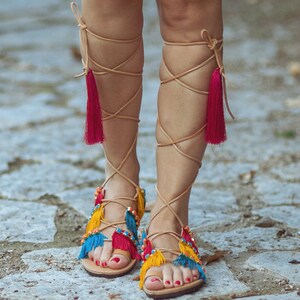 May include: A pair of tan leather sandals with colourful tassels. The sandals have multiple straps that wrap around the ankles and legs. The tassels are red, yellow, and blue.