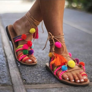 May include: A pair of brown leather sandals with colourful pom-poms and tassels. The sandals have a lace-up design and are decorated with a variety of colours, including pink, yellow, orange, and blue.