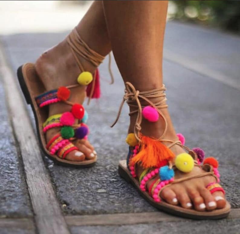May include: A pair of brown leather sandals with colourful pom-poms and tassels. The sandals have a lace-up design and are decorated with a variety of colours, including pink, yellow, orange, and blue.