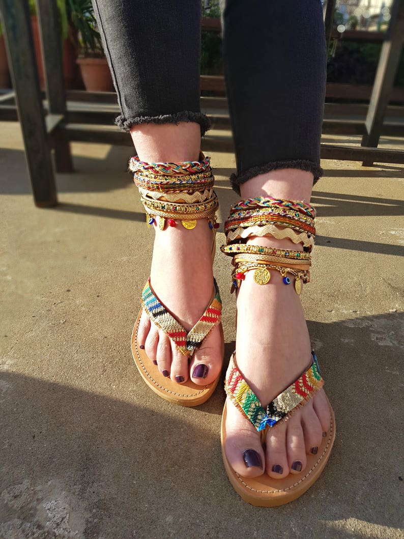 May include: A pair of brown leather sandals with colourful woven straps and gold accents. The sandals are adorned with multiple gold anklets with charms, including evil eye beads.