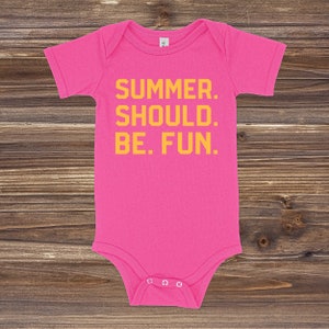 May include: A bright pink baby onesie with the text "SUMMER. SHOULD. BE. FUN." in yellow block letters. The onesie has short sleeves and snaps at the bottom. The background is a wooden surface.
