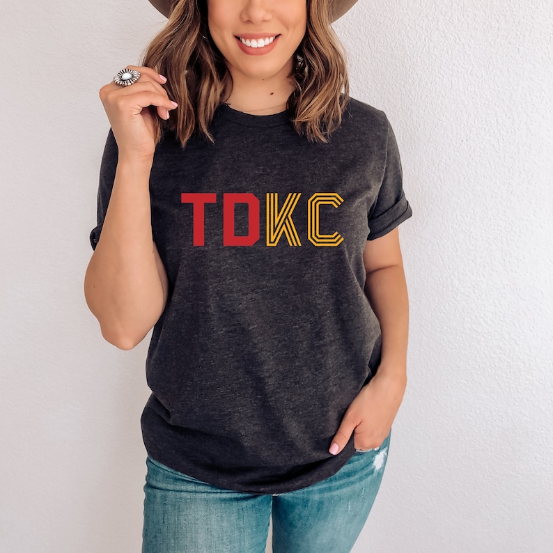 Op de afbeelding: Een donkergrijs T-shirt met een rode "TDKC" print op de voorkant. De letters zijn geel omrand.