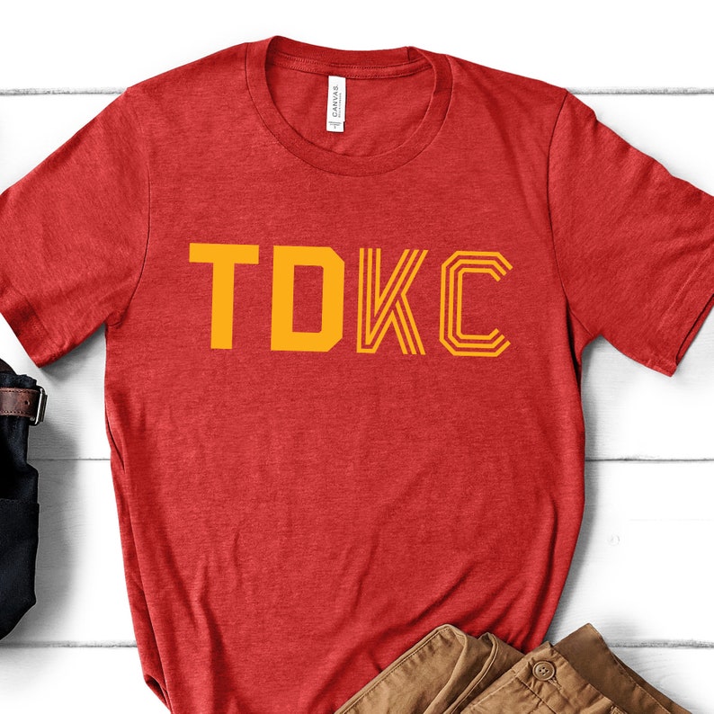 Op de afbeelding: Rood T-shirt met de tekst "TDKC" in geel.