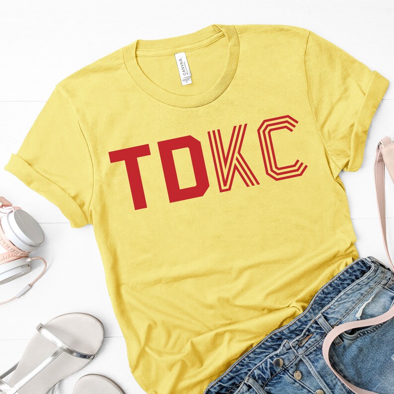 Op de afbeelding: Geel T-shirt met korte mouwen met de tekst "TDKC" in rood.