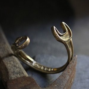 Könnte beinhalten: Ein goldfarbenes Metallarmband in Form eines Schraubenschlüssels. Das Armband liegt auf einem abgenutzten Hammer.