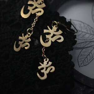 May include: Gold earrings with a chain and multiple Om symbols hanging from them. The earrings are on a black fabric background.
