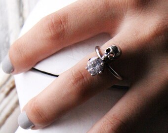 Puede incluir: Un anillo de plata con una calavera y una piedra preciosa transparente. El anillo se lleva en un dedo que sostiene una escultura de calavera blanca.
