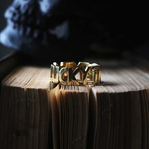 Op de afbeelding: Een gouden ring met het woord "LUCKY" gespeld in reliëfletters. De ring ligt op een stapel oude, versleten boekpagina's.