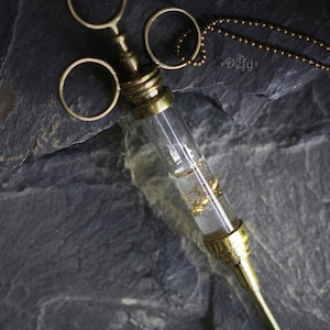May include: A gold-toned necklace with a glass vial pendant containing small gold beads. The vial is attached to a brass syringe-like structure with three rings. The necklace is on a dark gray background.
