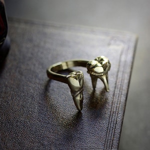 Puede incluir: Un anillo dorado con dos grandes detalles en forma de dientes. El anillo está sobre una superficie de cuero marrón oscuro.