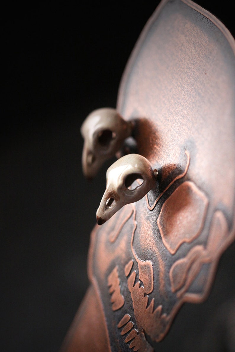 Puede incluir: Una escultura de calavera de cer&aacute;mica con un esmalte de cobre. La calavera tiene dos peque&ntilde;os cr&aacute;neos de p&aacute;jaros de cer&aacute;mica blanca unidos a la parte superior.