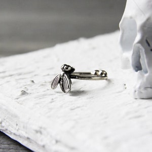 May include: A silver ring with a detailed fly design. The ring is on a white surface next to a white skull.