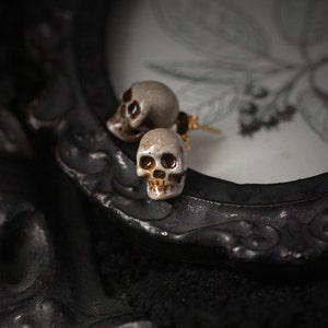 Puede incluir: Un par de pendientes en forma de calavera con detalles dorados. Los pendientes est&aacute;n hechos de cer&aacute;mica blanca y tienen un dise&ntilde;o detallado.