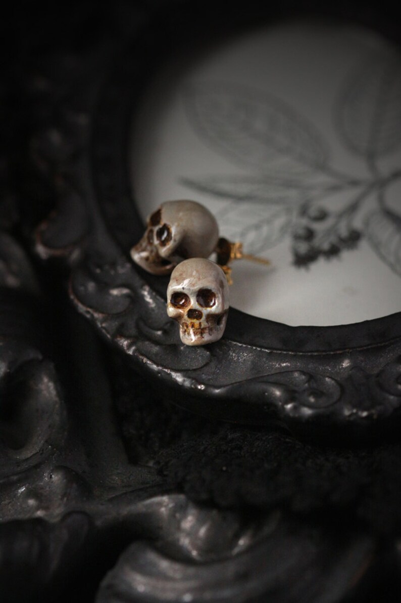 Puede incluir: Un par de pendientes en forma de calavera con detalles dorados. Los pendientes est&aacute;n hechos de cer&aacute;mica blanca y tienen un dise&ntilde;o detallado.