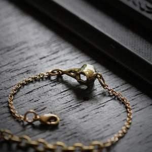 May include: A gold chain bracelet with a small bird skull charm. The bracelet is on a dark wooden surface.