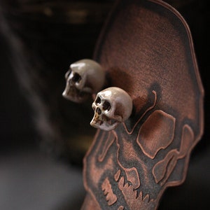 Puede incluir: Dos peque&ntilde;os pendientes de calavera de cer&aacute;mica blanca en un expositor de calavera de cobre.