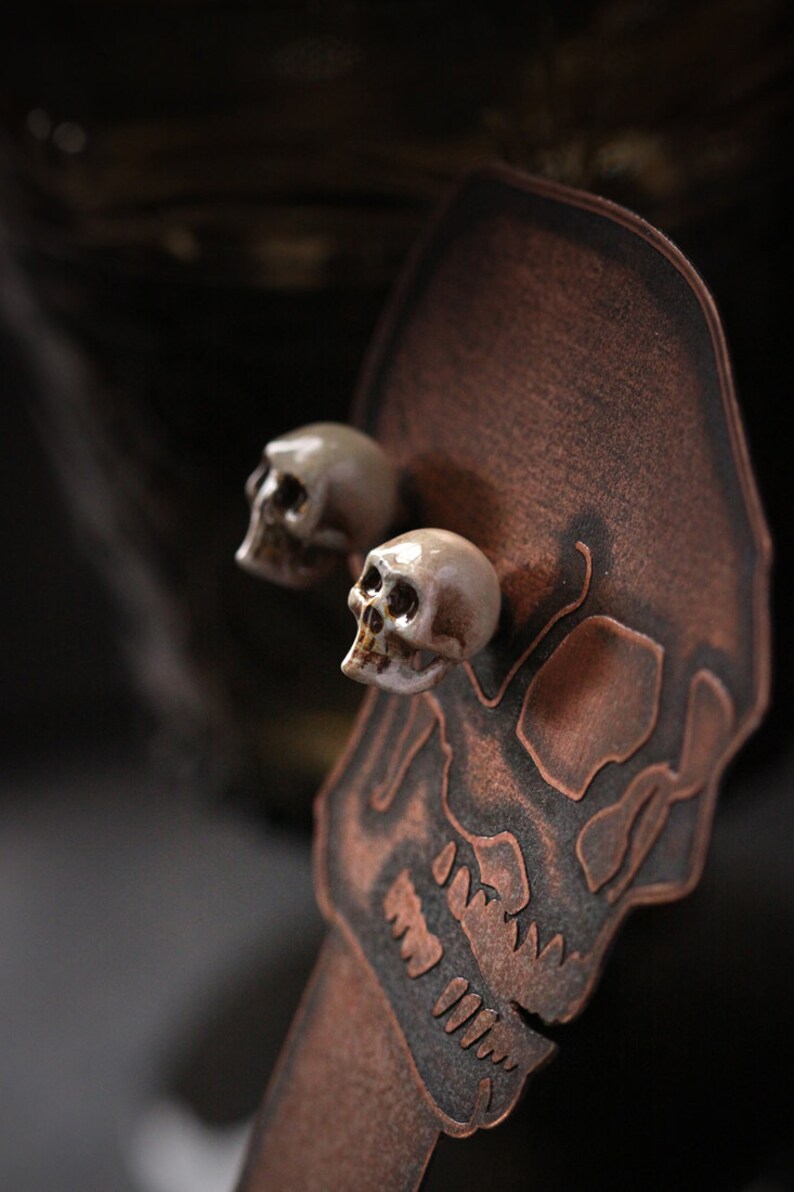 Puede incluir: Dos peque&ntilde;os pendientes de calavera de cer&aacute;mica blanca en un expositor de calavera de cobre.