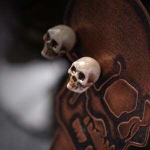 Puede incluir: Dos peque&ntilde;os pendientes de cer&aacute;mica blanca en forma de calavera con cuencas de ojos negras. Los pendientes se muestran sobre un fondo de cuero marr&oacute;n con un dise&ntilde;o de calavera.