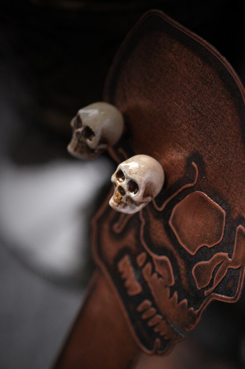 Puede incluir: Dos peque&ntilde;os pendientes de cer&aacute;mica blanca en forma de calavera con cuencas de ojos negras. Los pendientes se muestran sobre un fondo de cuero marr&oacute;n con un dise&ntilde;o de calavera.