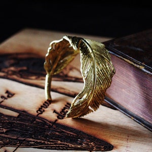 May include: A gold-coloured feather ring, with a detailed design, is resting on a wooden surface with an engraved design.