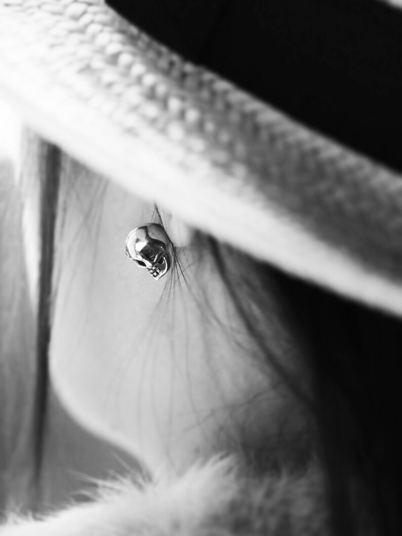 Puede incluir: Un primer plano de un pendiente de calavera de plata. El pendiente es peque&ntilde;o y delicado, y lo lleva una persona con el pelo largo.