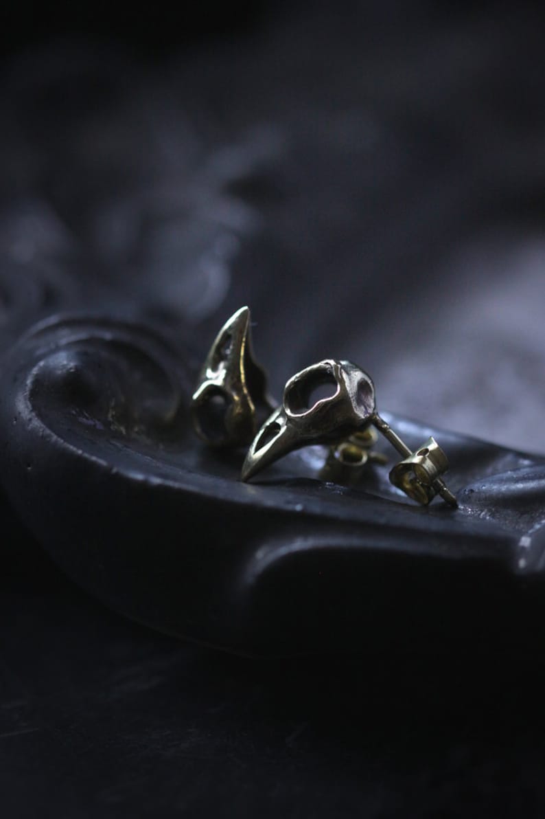 May include: A pair of silver skull-shaped earrings with a detailed design. The earrings are on a black surface.