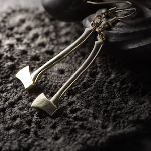May include: A pair of gold-toned earrings shaped like miniature axes. The earrings are hanging from a black surface.