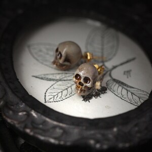 Puede incluir: Un par de pendientes en forma de calavera con detalles dorados. Los pendientes est&aacute;n sobre un fondo blanco con un dise&ntilde;o floral.