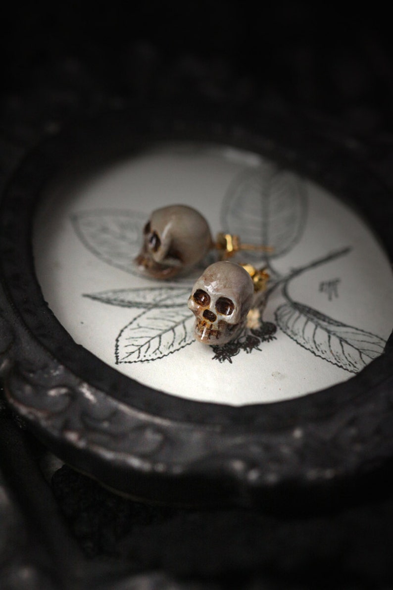 Puede incluir: Un par de pendientes en forma de calavera con detalles dorados. Los pendientes est&aacute;n sobre un fondo blanco con un dise&ntilde;o floral.