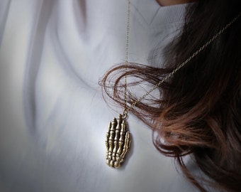 May include: A gold skeleton hand pendant necklace on a delicate chain. The pendant is detailed and intricate, with each bone of the hand clearly visible.