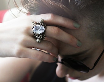 Puede incluir: Un anillo plateado con una gema grande, ovalada y transparente. El anillo tiene un diseño detallado y ornamentado con detalles de calaveras. La mano de la persona descansa sobre su frente, con uñas pintadas. El anillo es el foco.