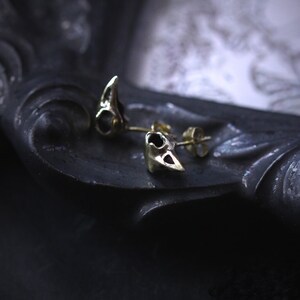 May include: A pair of gold-toned earrings shaped like small bird skulls. The earrings are on a dark surface.