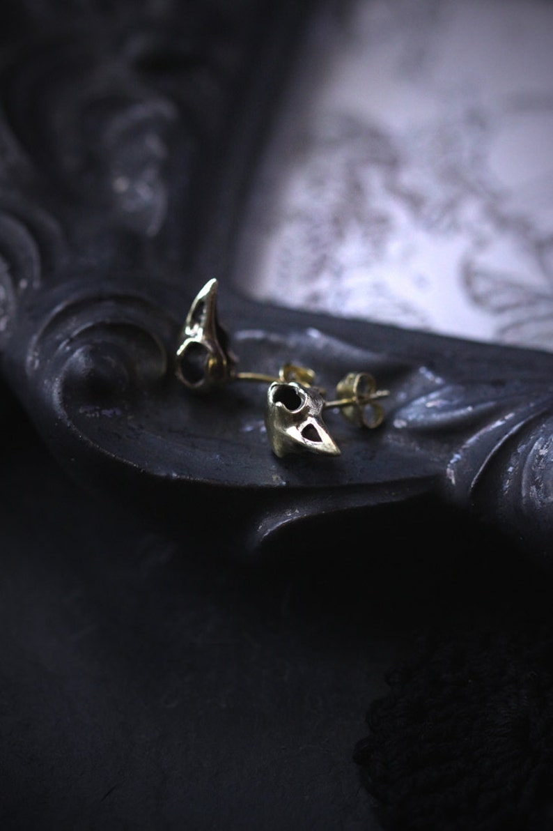 May include: A pair of gold-toned earrings shaped like small bird skulls. The earrings are on a dark surface.