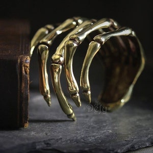 Op de afbeelding: Een goudkleurige manchetarmband in de vorm van een skelet hand. De armband is gemaakt van metaal en heeft een gedetailleerd ontwerp van een menselijke hand. De armband ligt op een donkere ondergrond met een boek op de achtergrond.