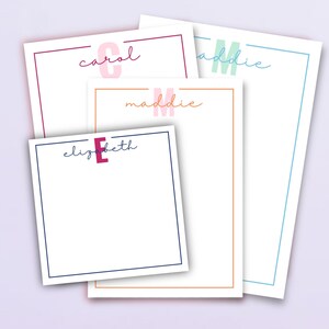 May include: A selection of personalised notepads featuring names in cursive writing. Each notepad has a coloured border and a matching initial. The names include Carol, Maddie, and Elizabeth. The notepads are white with coloured borders.