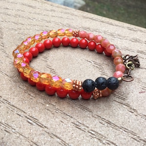 Puede incluir: Un brazalete de capas con cuentas rojas, naranjas y negras. El brazalete está hecho con cuentas de lava, vidrio y cobre.