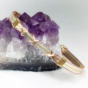 Könnte beinhalten: Ein Armband aus Silber-, Gold- und Kupferdraht mit geflochtenem Design. Das Armband liegt auf einem violetten Amethyst-Kristallcluster.
