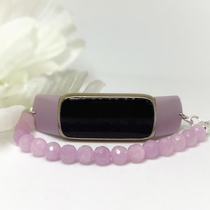 Puede incluir: Un rastreador de fitness lila con una pantalla negra y un marco de metal plateado. Una pulsera de cuentas lila a juego está adjunta. El fondo incluye una flor blanca.