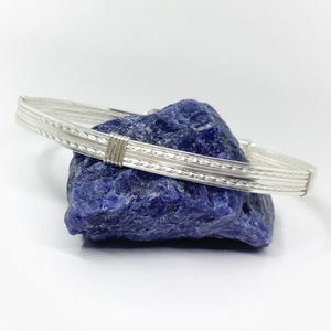May include: A silver-colored bangle bracelet featuring three parallel bands, with the outer two bands displaying a twisted rope texture and the center band being smooth. A small, rectangular silver-colored wire wrap secures the three bands together.
