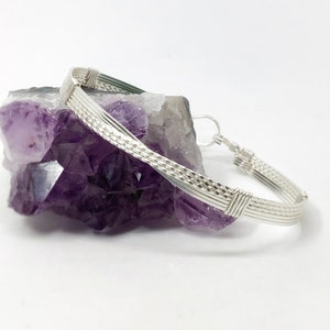 May include: A silver-toned bracelet with a woven design and a hook clasp. The bracelet is displayed on a purple crystal formation against a white background. The bracelet's design includes multiple parallel wires.
