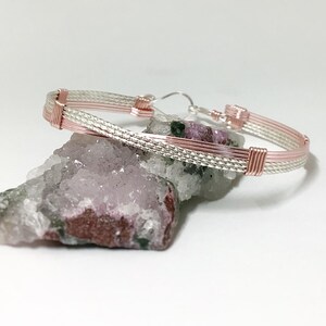 May include: A silver and rose gold wire bracelet with a braided design. The bracelet is resting on a pink and green crystal.
