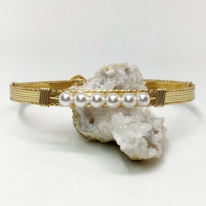 Puede incluir: Un brazalete de alambre dorado envuelto con una fila de perlas blancas.